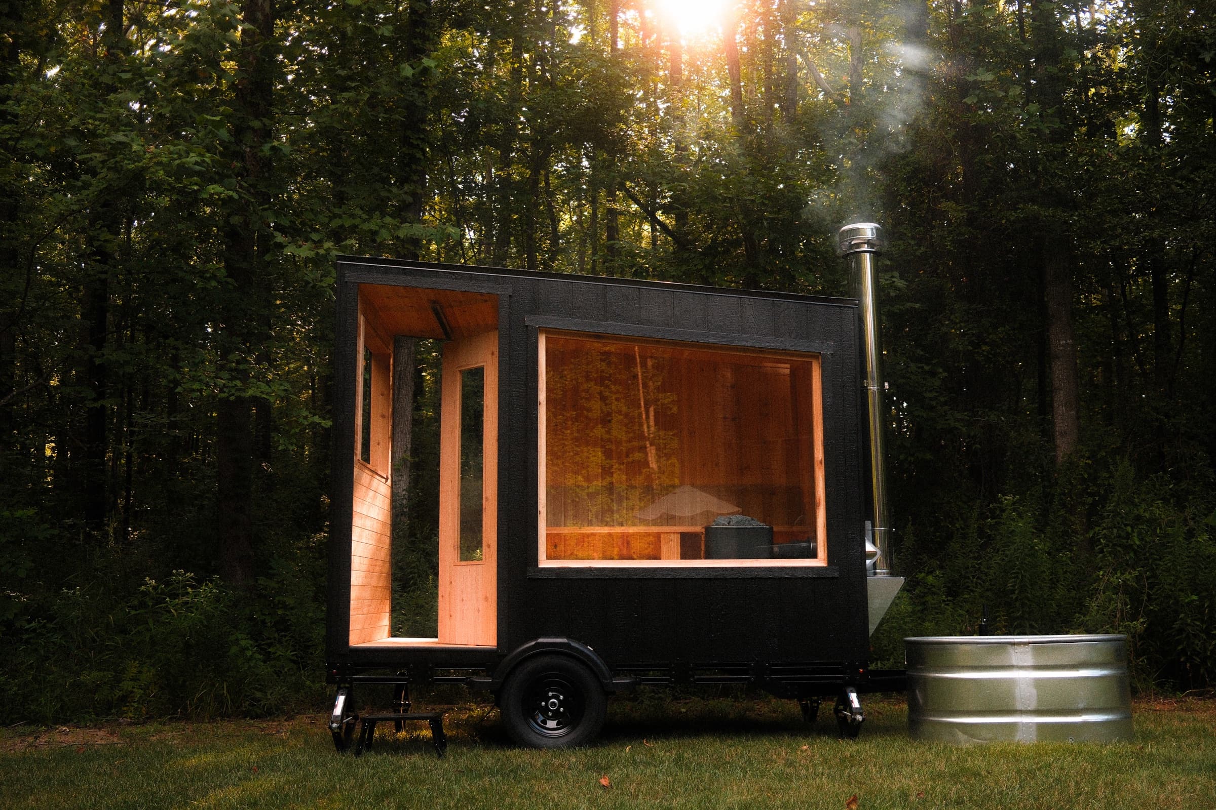 A wood-fired sauna on a trailer at the edge of a forest, two people sitting inside, with a cold plunge tub alongside.